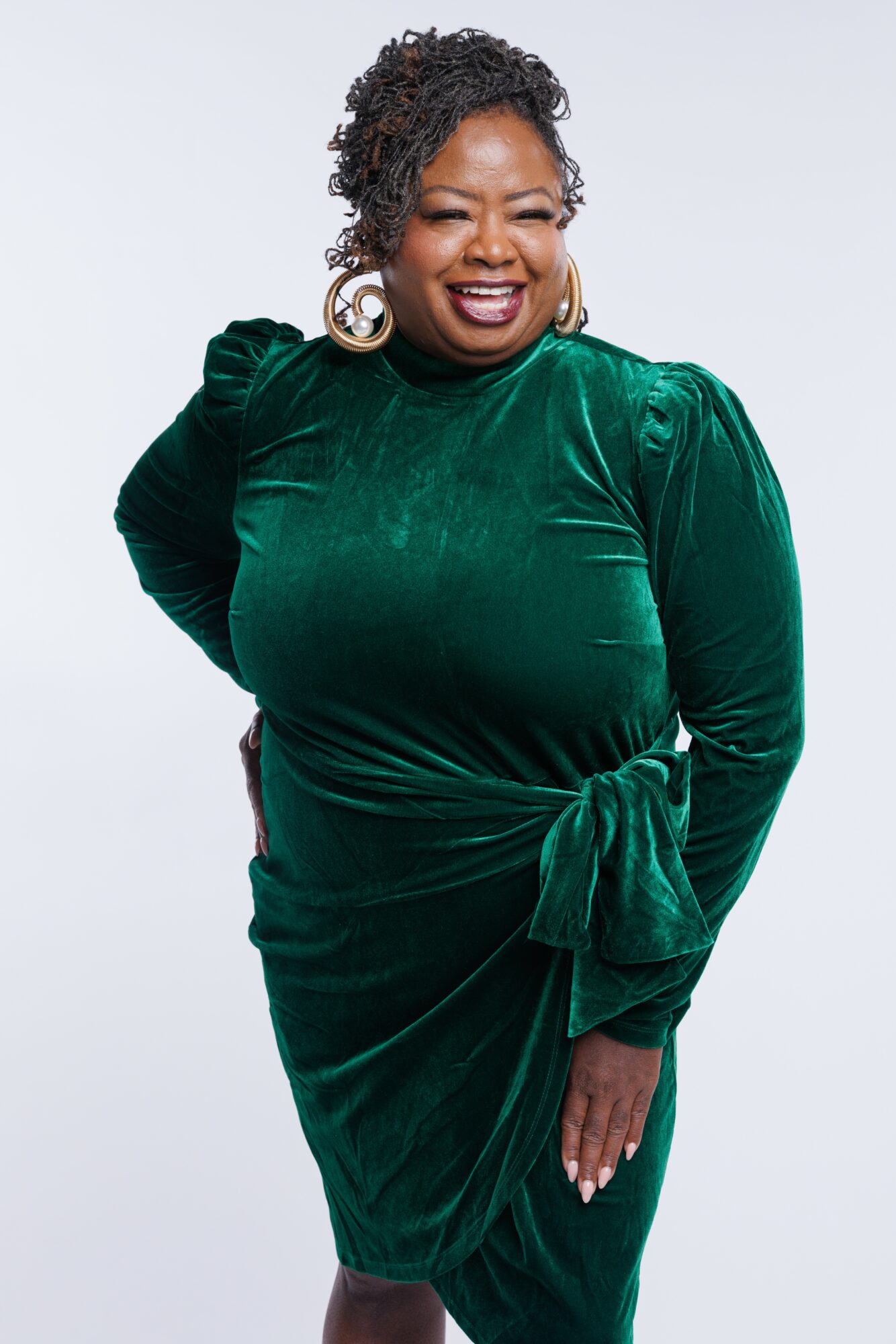 Smiling woman in green velvet dress with bow, curly hair, earrings, standing against white background.