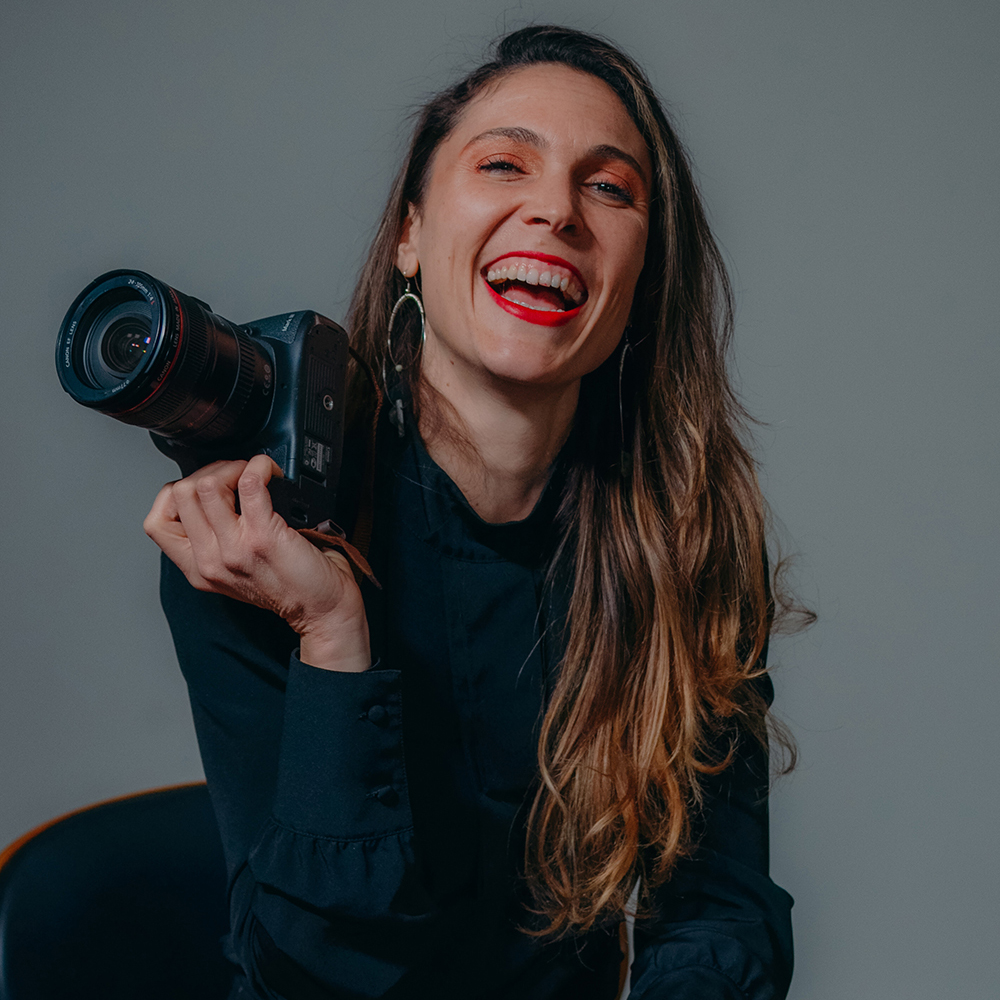 Woman with long hair smiling, holding a camera, wearing a dark top, against a plain background.
