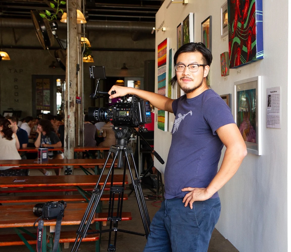 Man with glasses standing next to a camera on a tripod in an art gallery, with colorful artwork on the wall behind him.