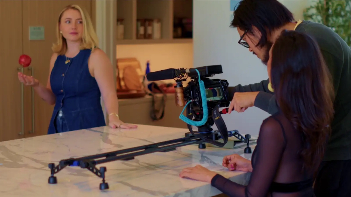 Two women and a man operate a camera on a track, with a woman holding a flower in a room, another woman sitting nearby.