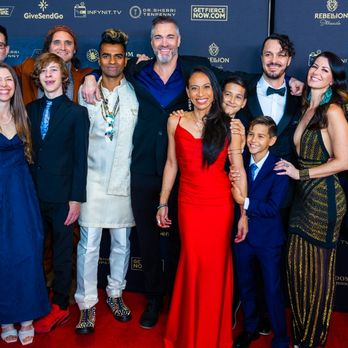 Group of people dressed in formal attire, smiling at an event with a blue backdrop. One woman in a red dress stands in the center.