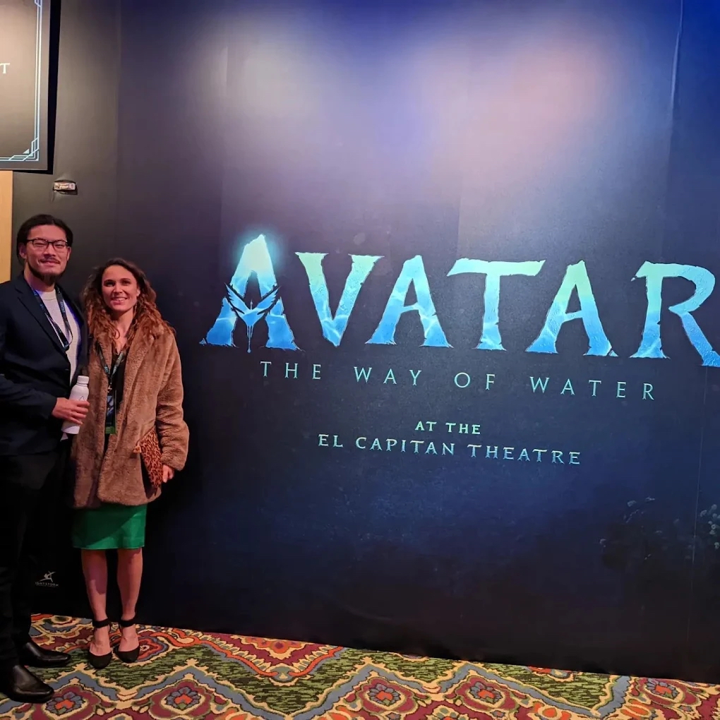 Two people standing in front of a large blue poster for the movie Avatar at El Capitan Theatre, with a patterned carpet below.