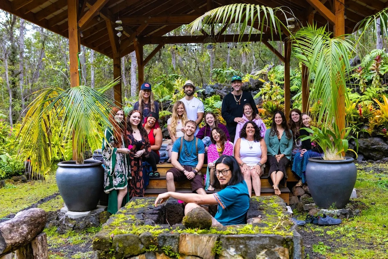 Group of people sitting and standing under a wooden shelter surrounded by lush greenery and large potted plants.
