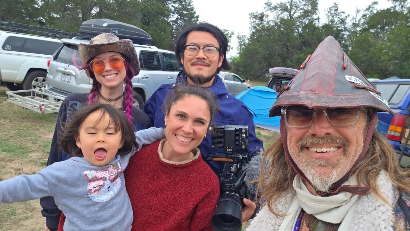 Group of five people outdoors, smiling, with trees and parked cars in background.