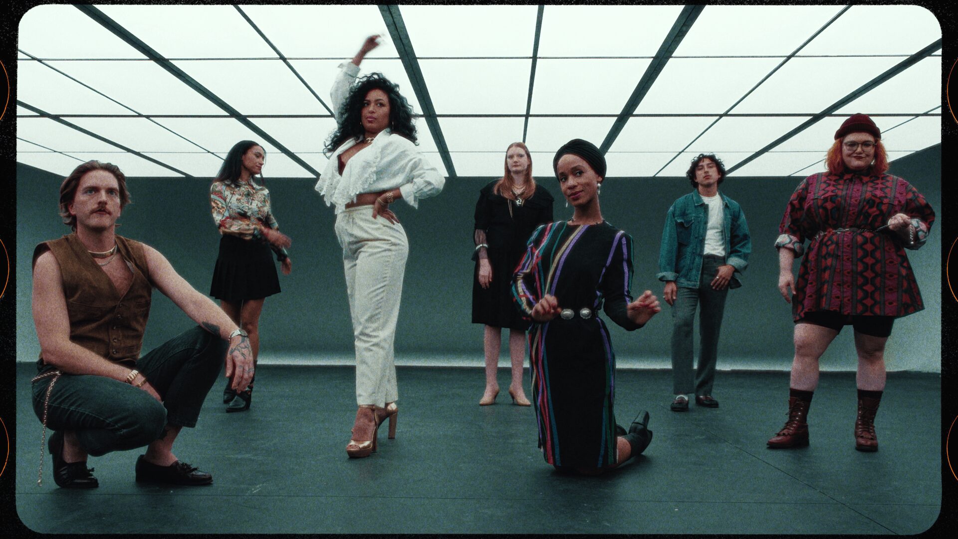 Seven women in a room with a grid ceiling, some standing and one kneeling, posing confidently.