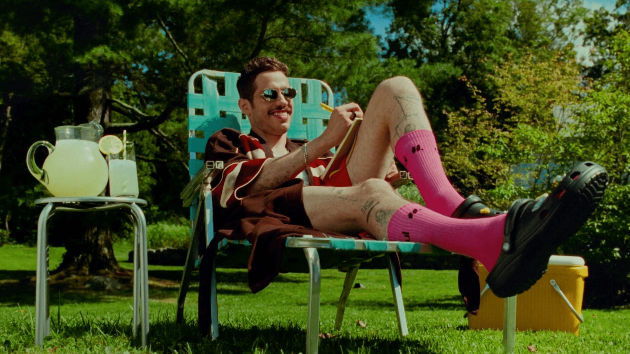 Person relaxing outdoors on a lounge chair, wearing sunglasses, pink socks, and smiling, with trees and blue sky in background.
