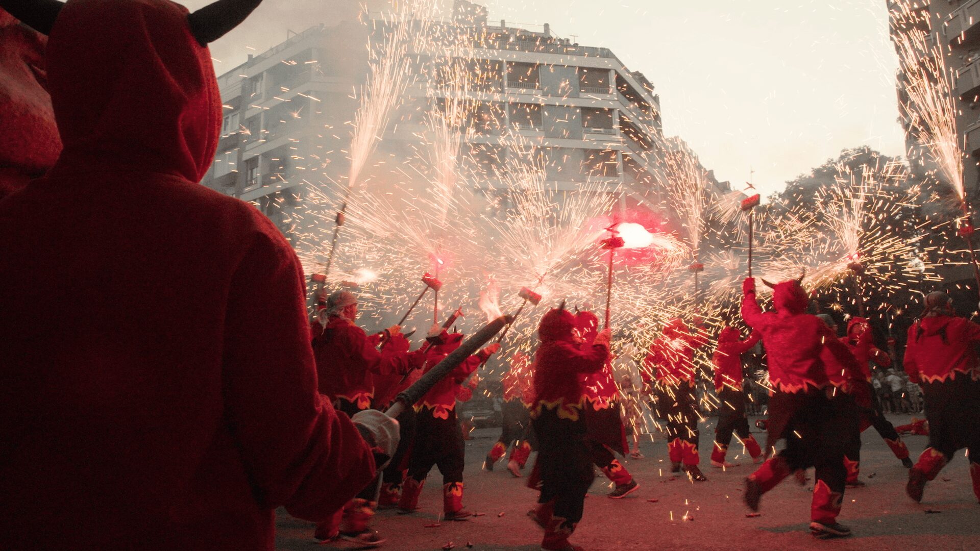 Group of people celebrating with fireworks and confetti in a city street, some wearing red costumes or masks.