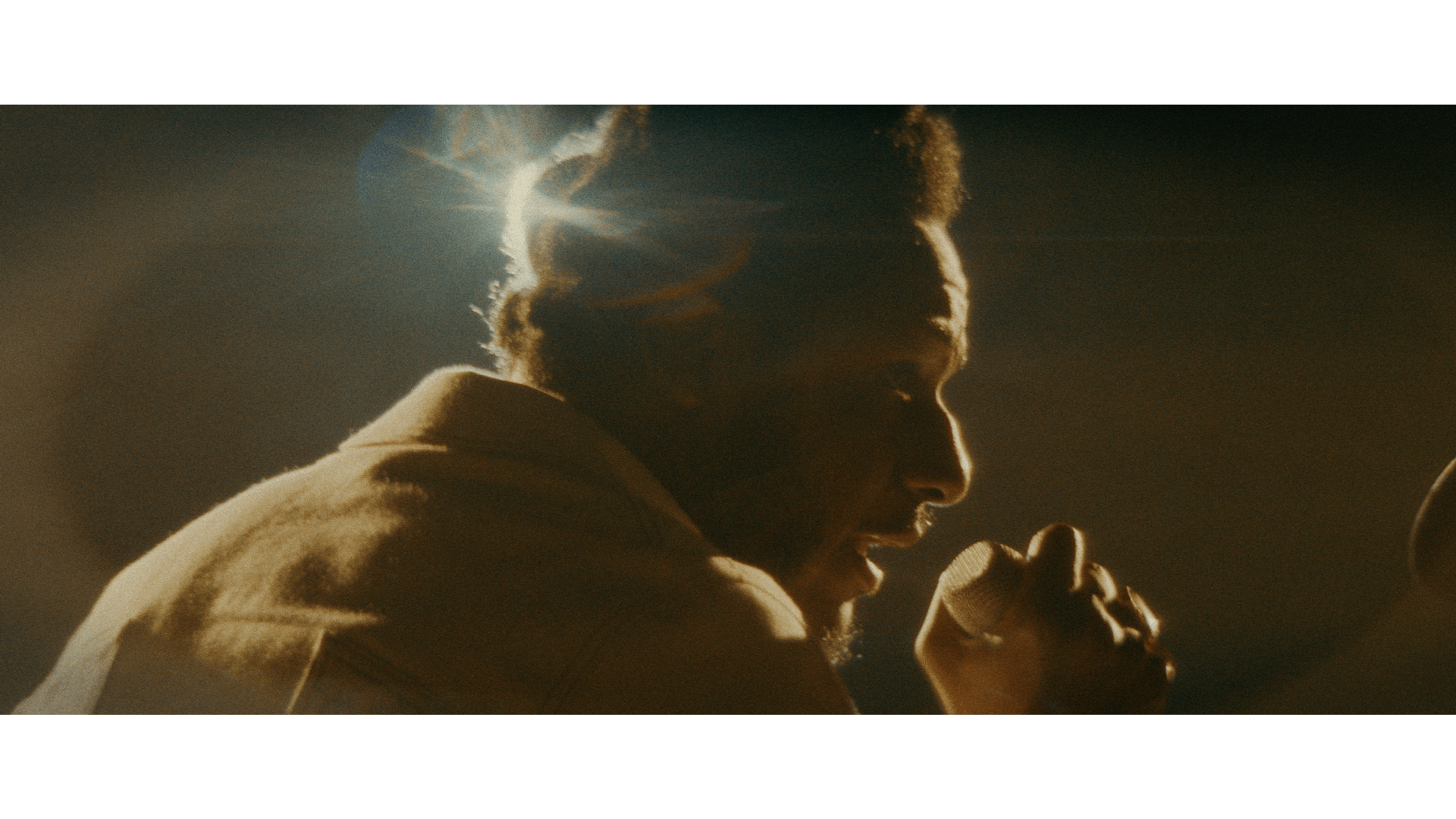 Person with curly hair in profile, illuminated by sunlight, appears to be speaking or singing, with a hand near their face.