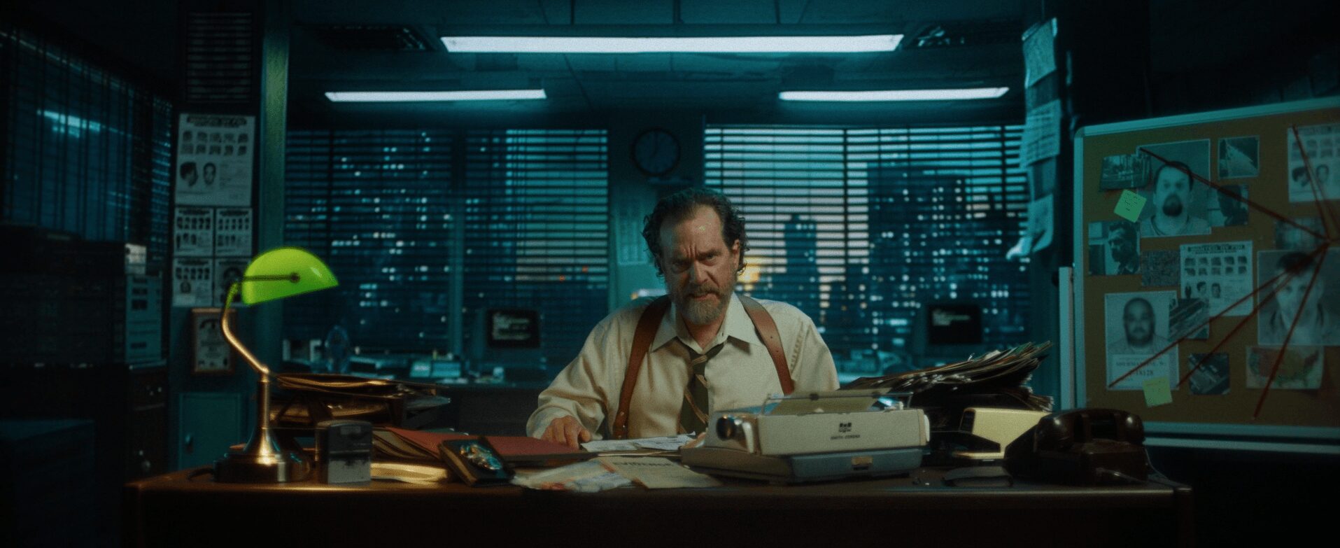 Man sitting at a cluttered desk in a dark office with city skyline view, illuminated by desk lamp.