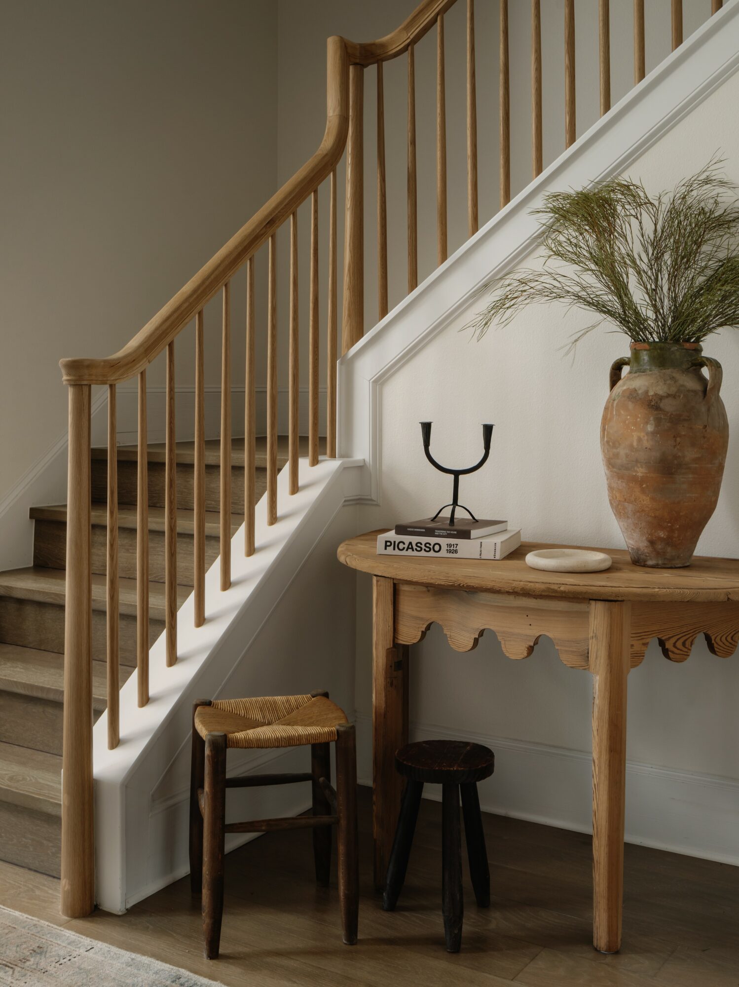 Staircase with wooden railing, small wooden table with a large vase and plants, two stools, and a book on the table.