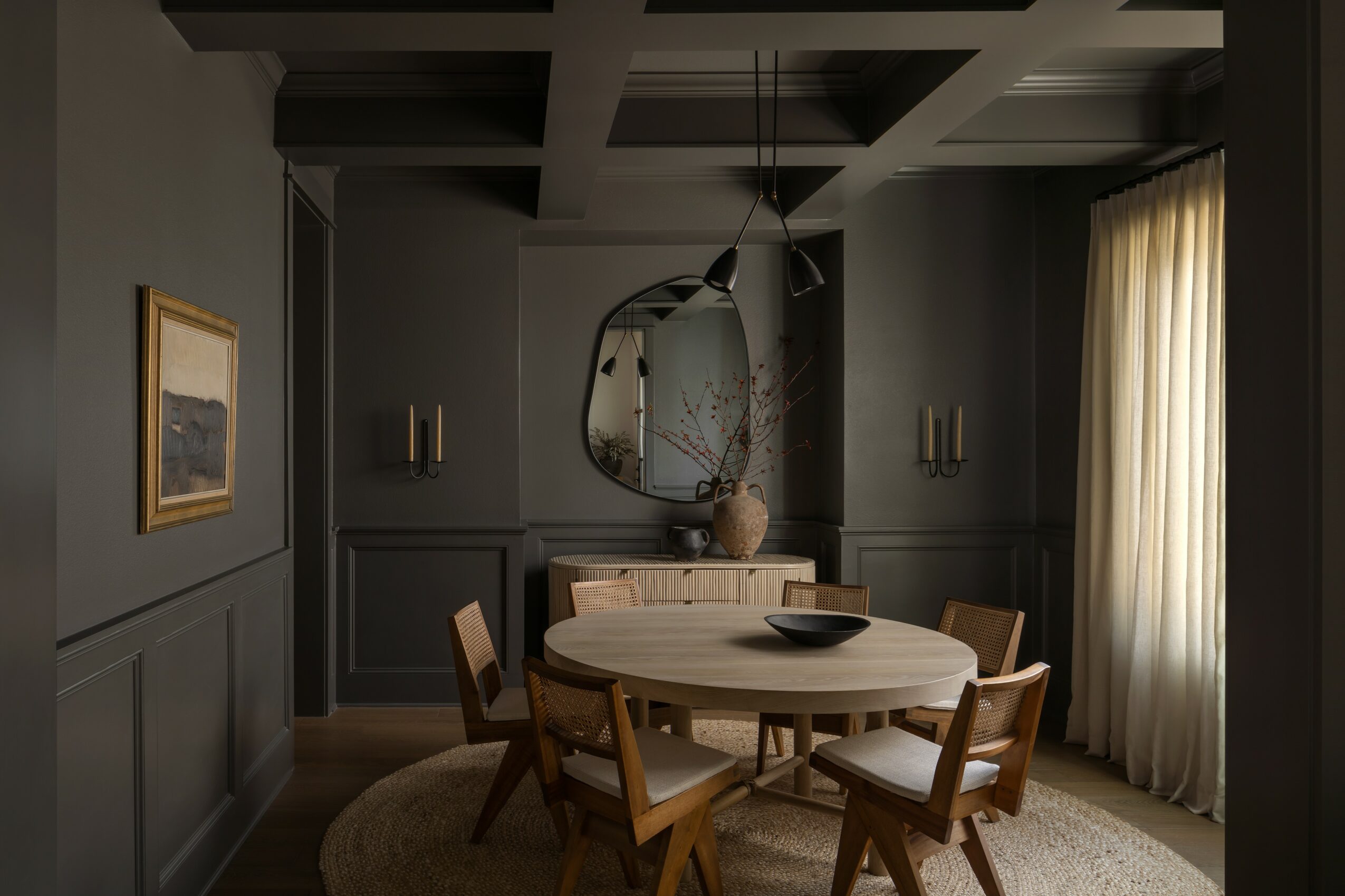 Dining room with round table, six chairs, mirror, wall art, and window with curtains, in a dimly lit space.