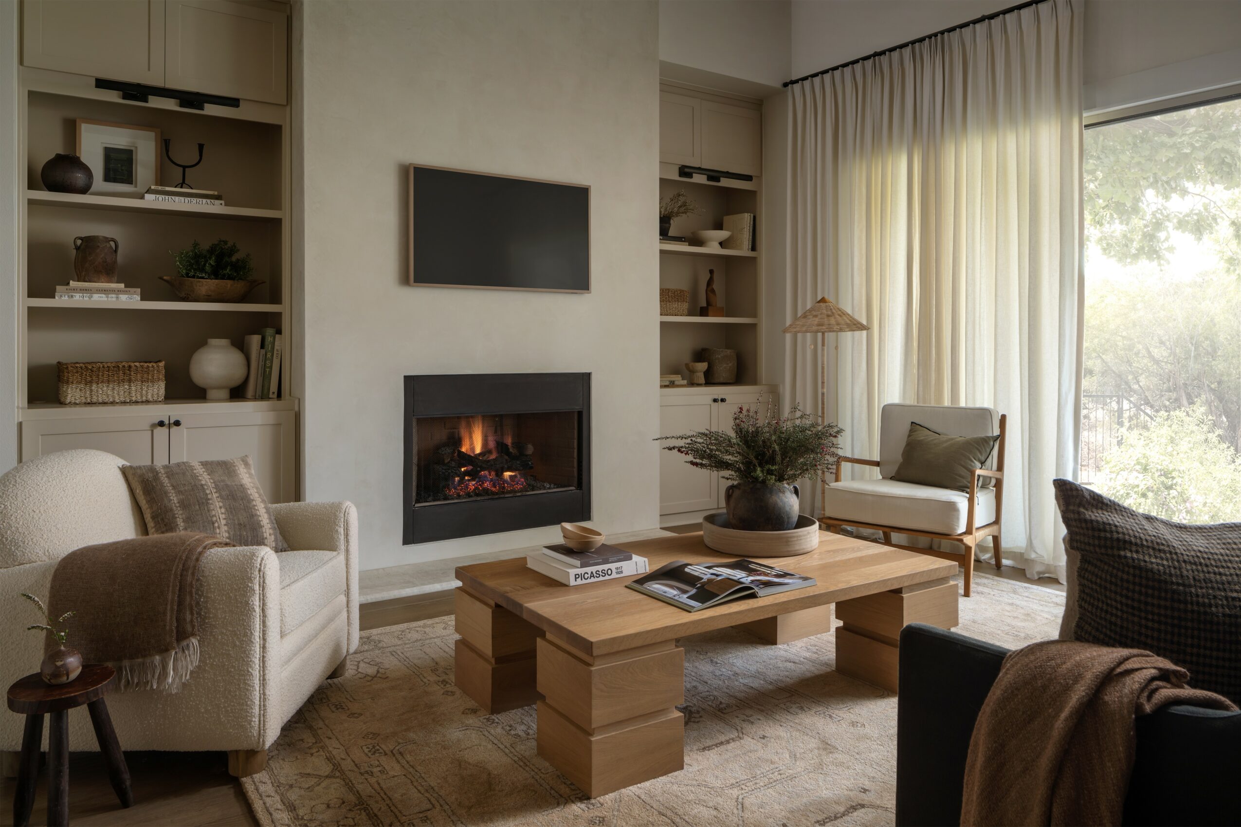 Living room with fireplace, TV, sofa, armchair, coffee table, and large window with curtains, sunlight outside.