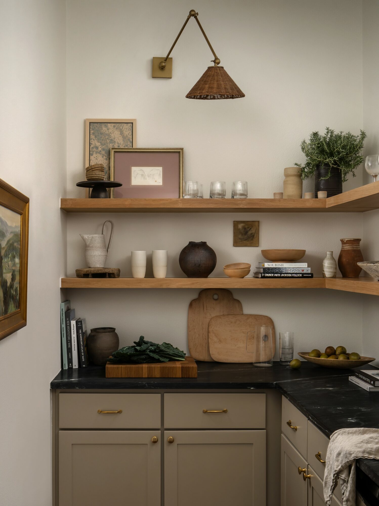 Kitchen shelf with decorative items, books, and plants, above beige cabinets with black countertop, wall with framed pictures and a hanging lamp.