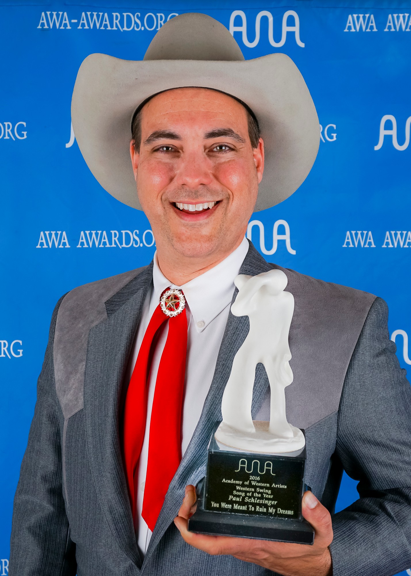 Man in cowboy hat holding award, smiling, blue background with AWA Awards logo, wearing gray suit and red tie.
