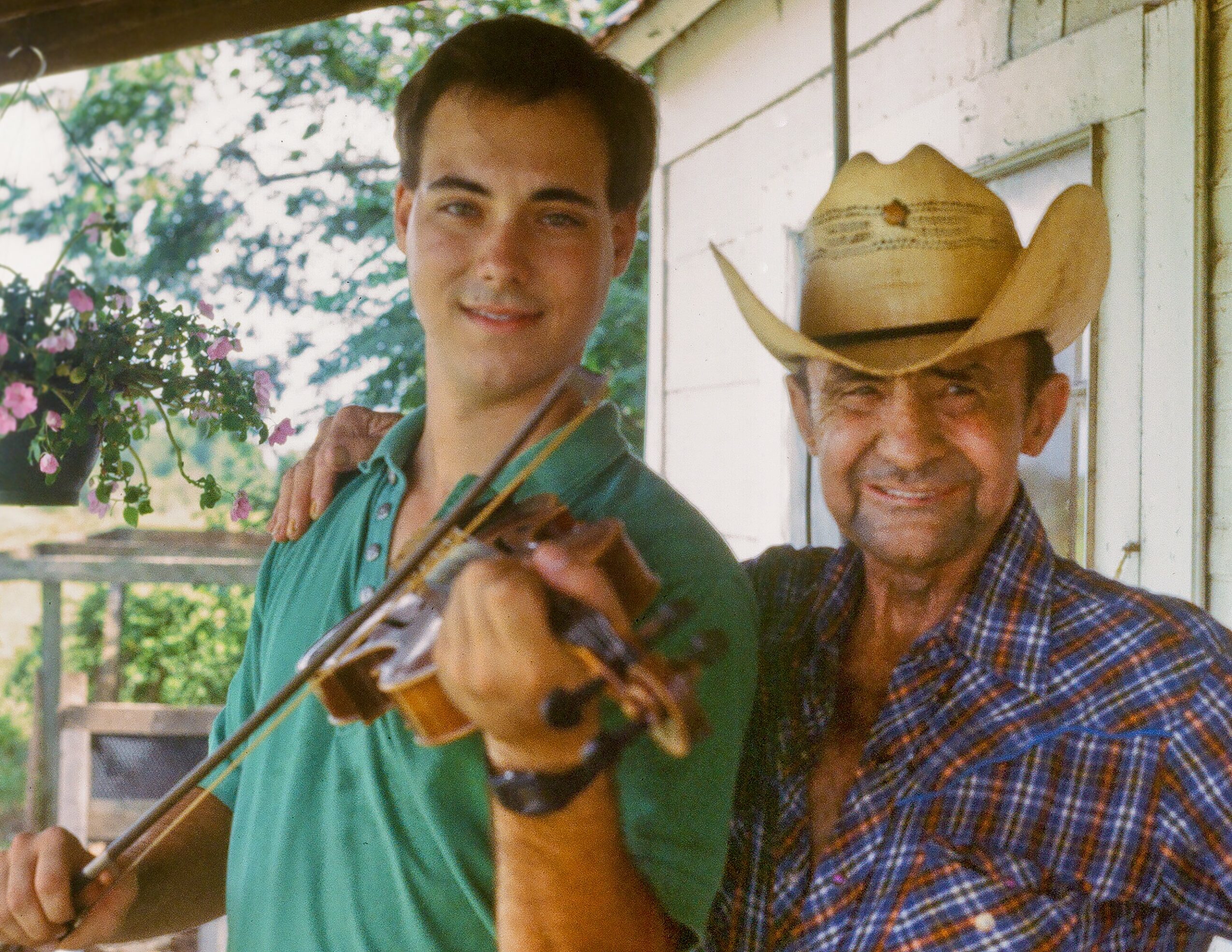 Two men outdoors, one young with a violin, the other older with a cowboy hat, smiling at the camera.