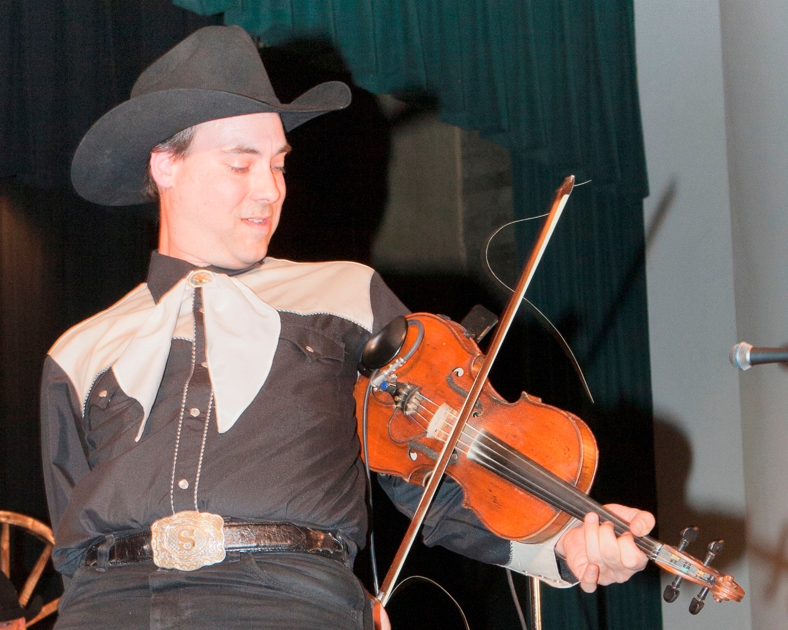 Person wearing cowboy hat playing a violin on stage, with a dark curtain in the background.