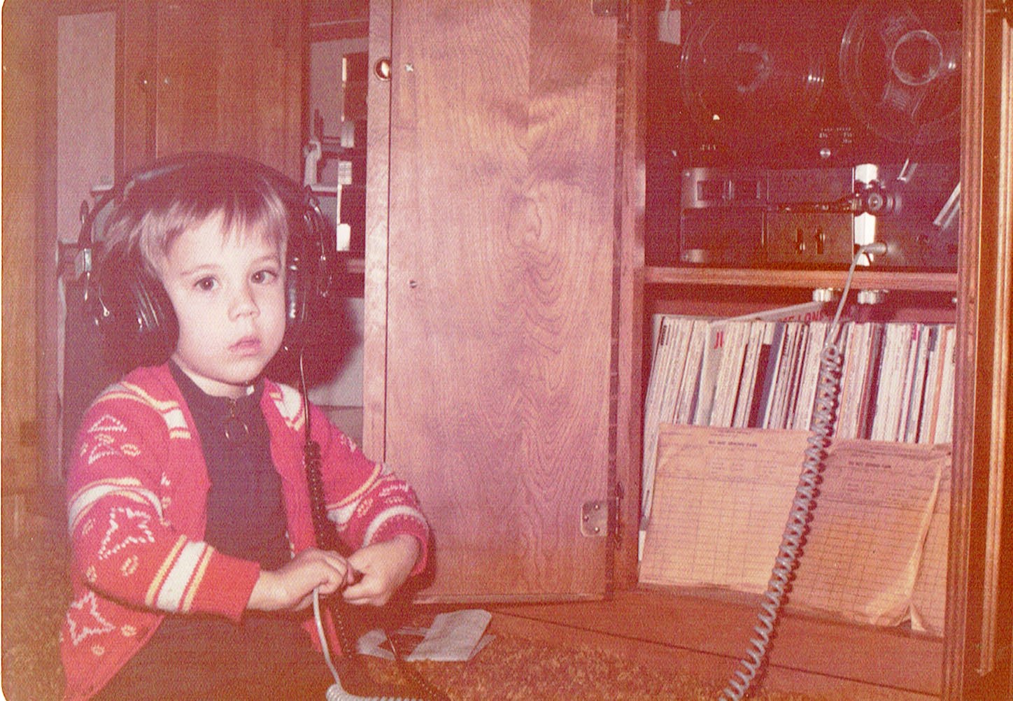 Young girl with headphones sitting at a desk with a telephone and records, wooden furniture in background.