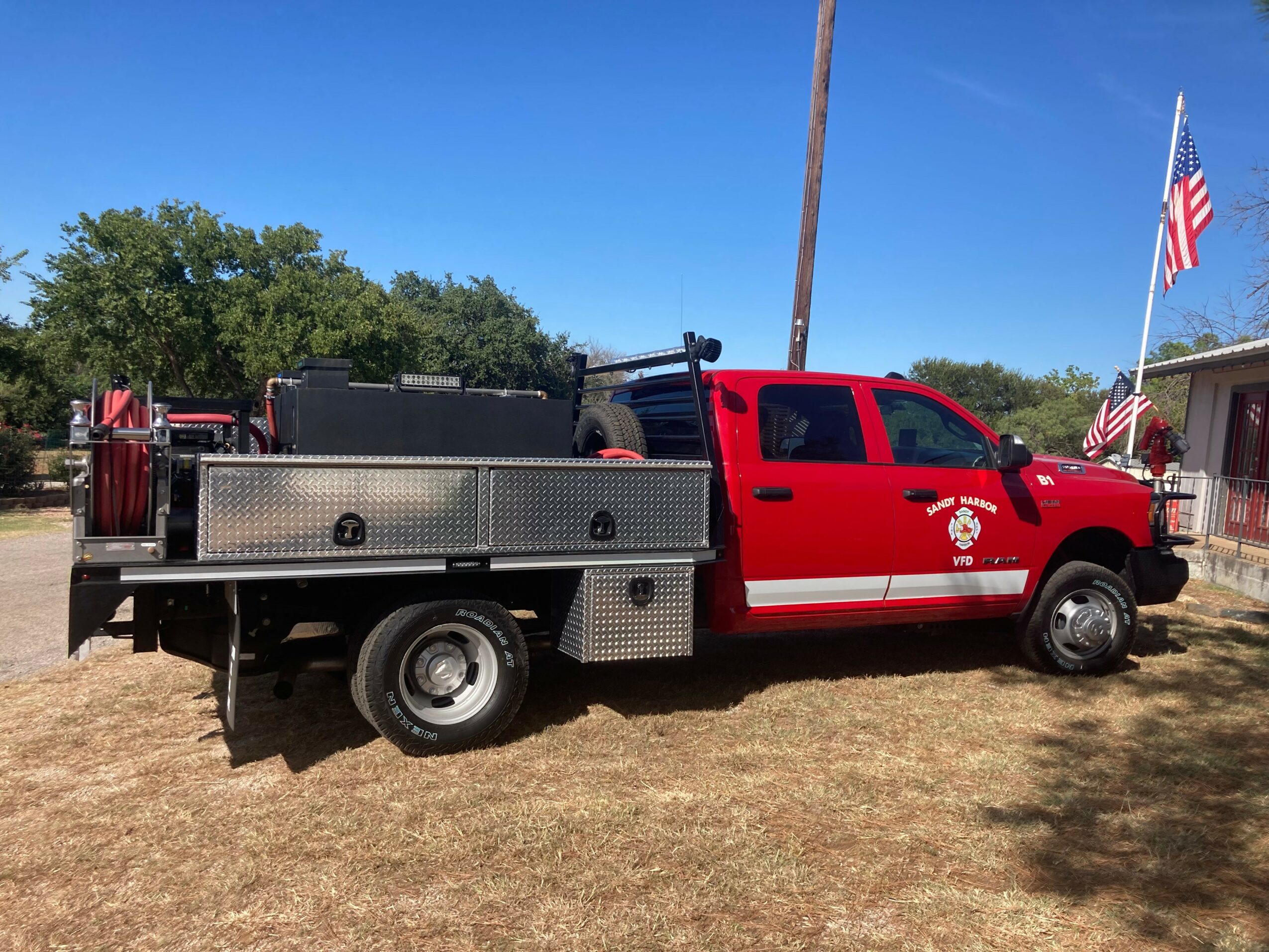 Red fire truck with equipment parked on grass, American flags in background, clear blue sky, trees nearby.