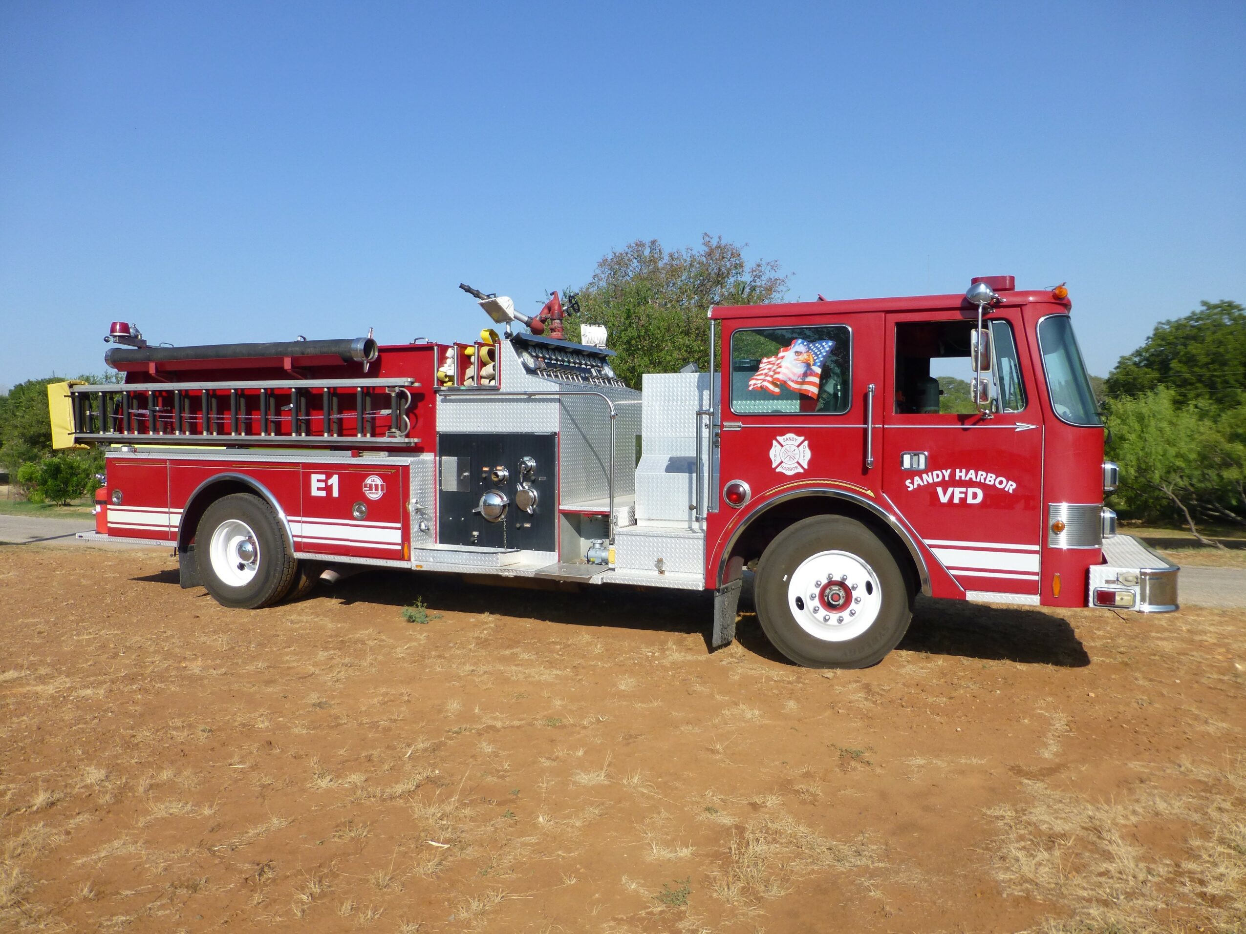 Red fire truck parked on dirt ground with trees and blue sky in background.
