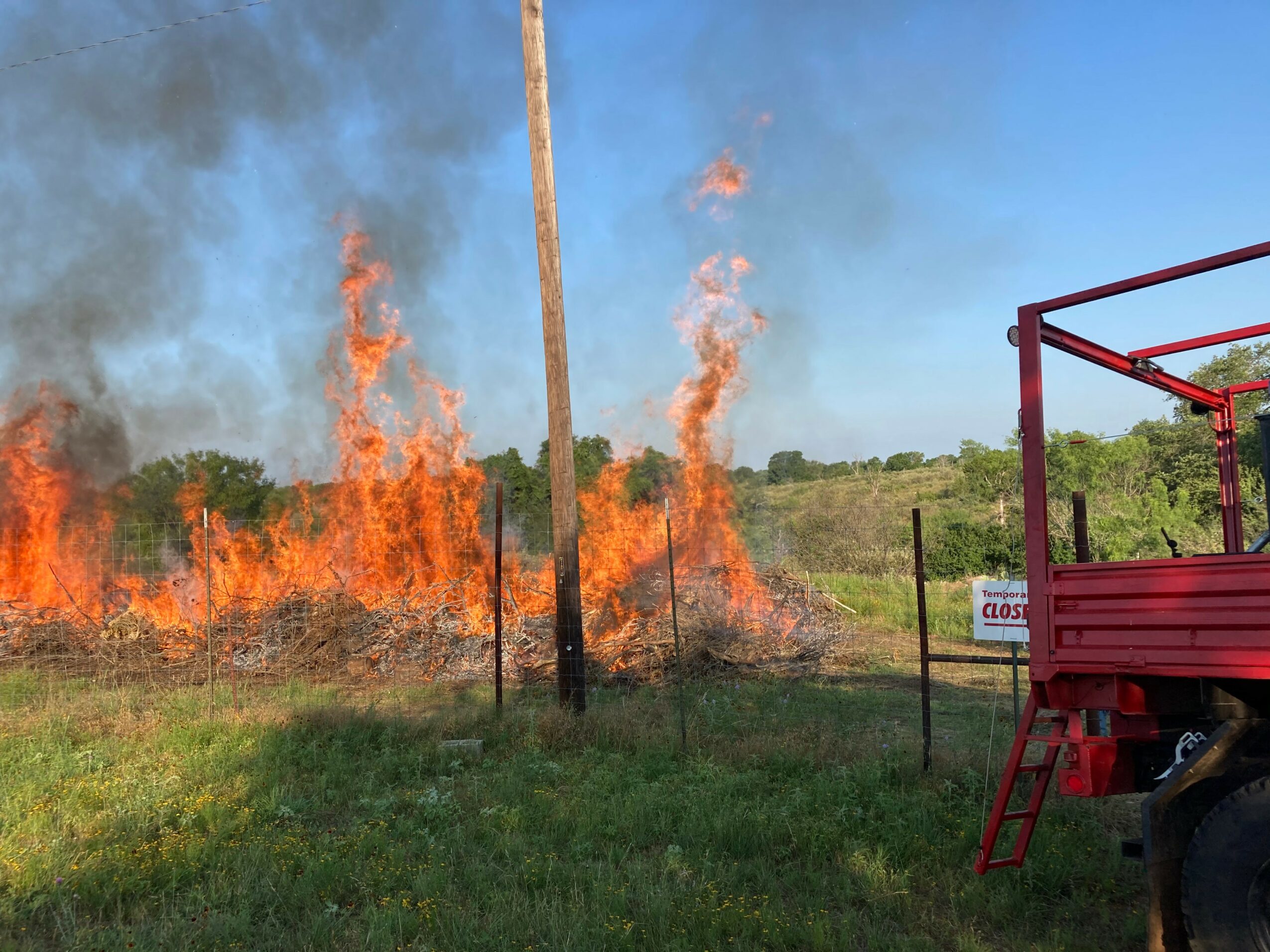 Fire burning in a field with smoke rising, a utility pole, and a red vehicle on the right side.