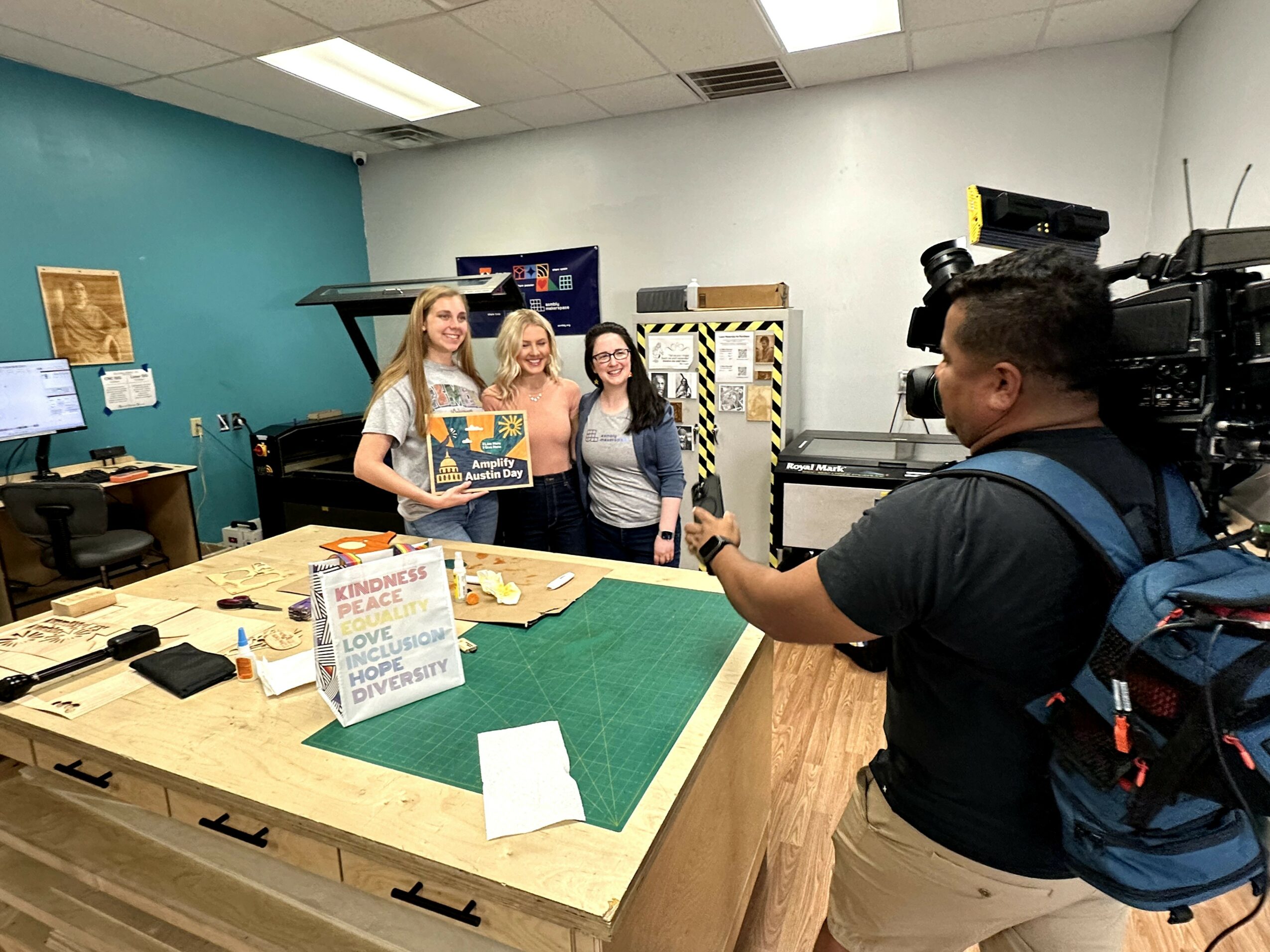 Three women stand behind a table with a green surface, being filmed by a cameraman in a room with blue and white walls.