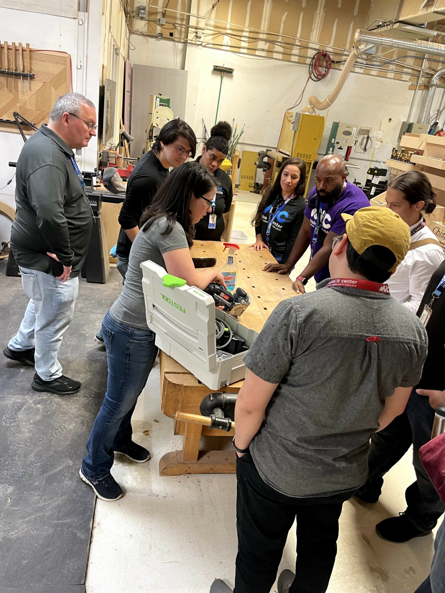 Group of people gathered around a workbench in a workshop, observing a demonstration or activity.