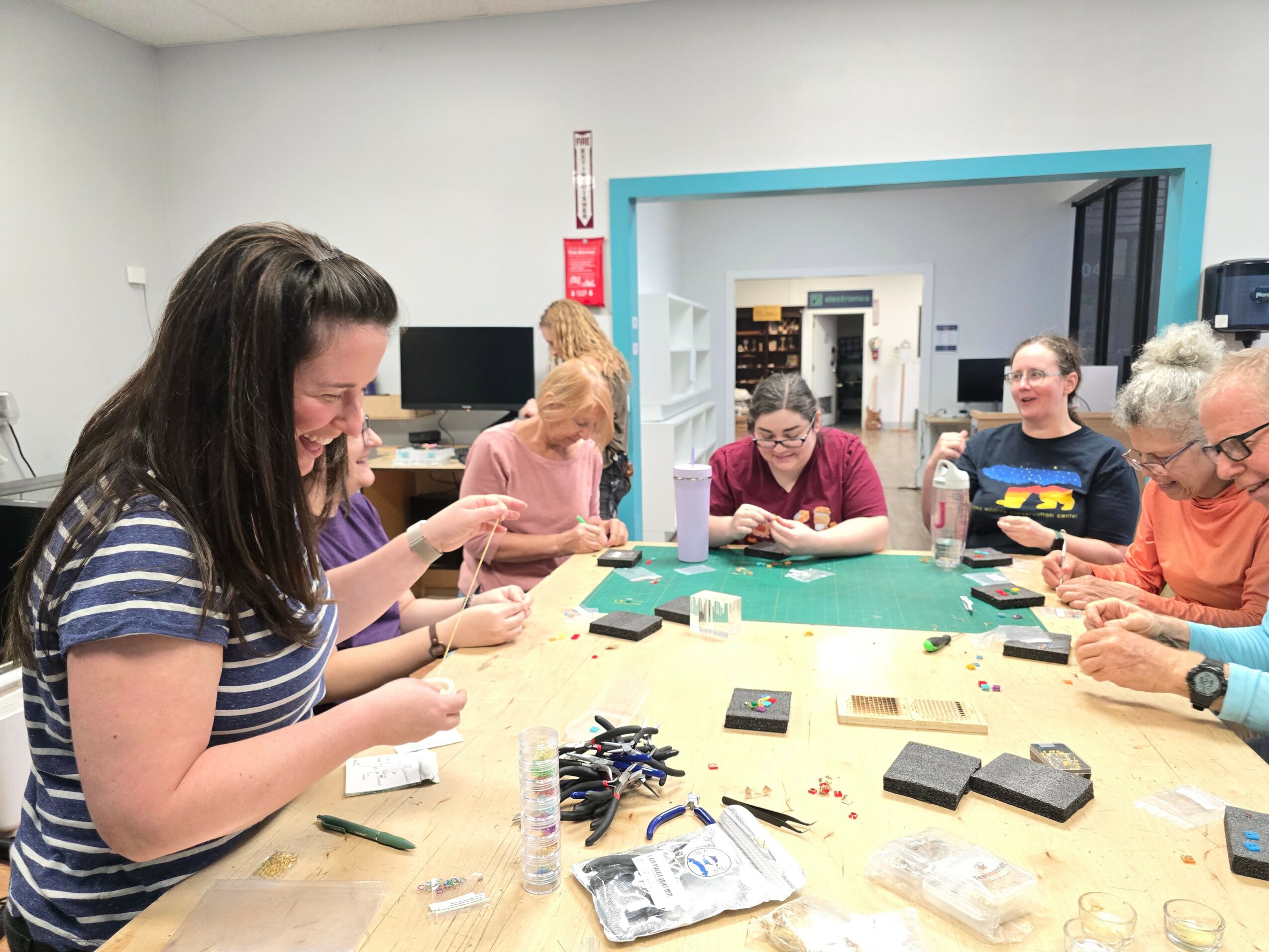 Group of six women sitting around a table working on electronic devices in a room with a blue doorway, some smiling and focused.