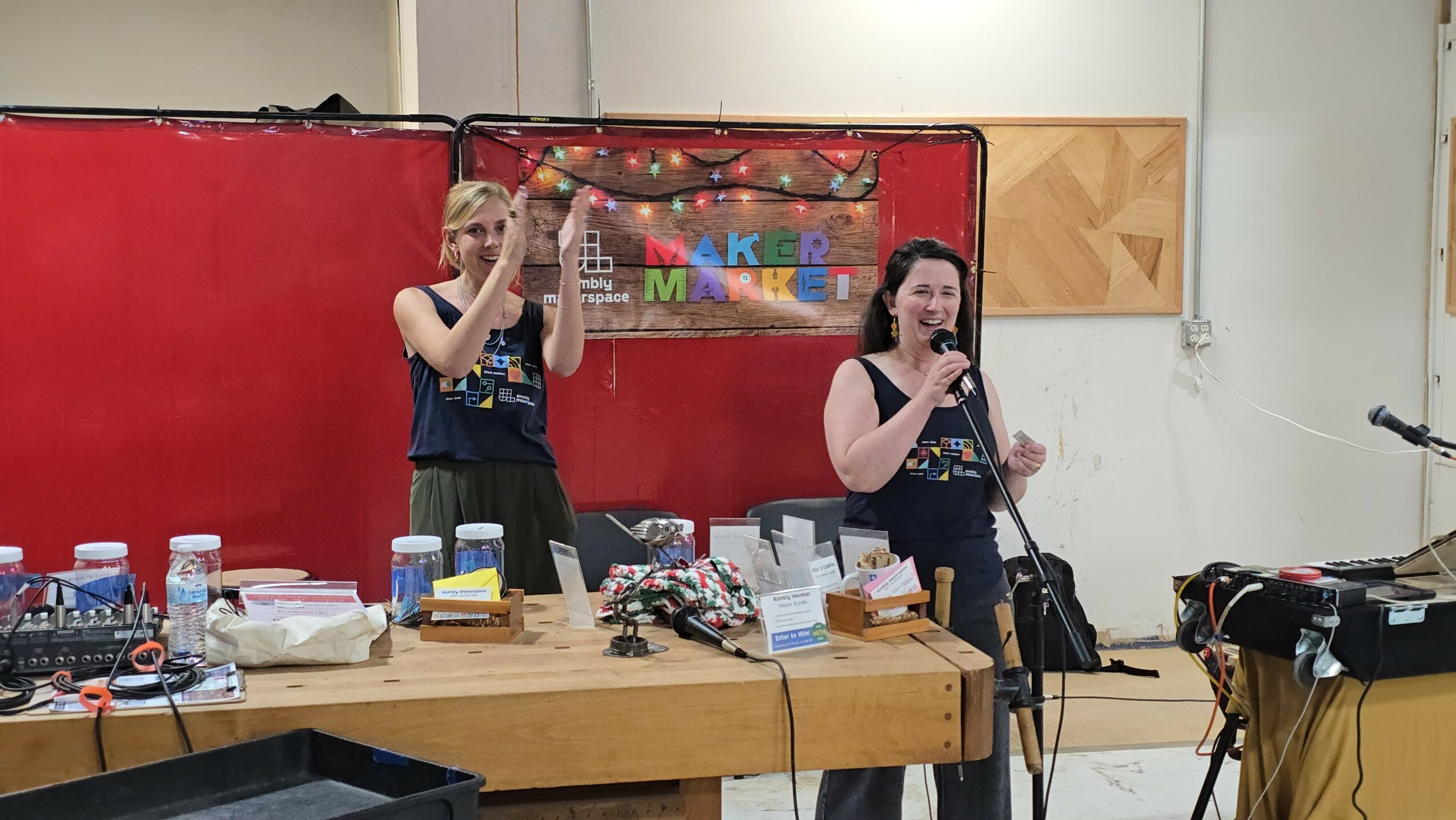 Two women singing into microphones in a room with a red and white background, a table with drinks and equipment, and a keyboard.
