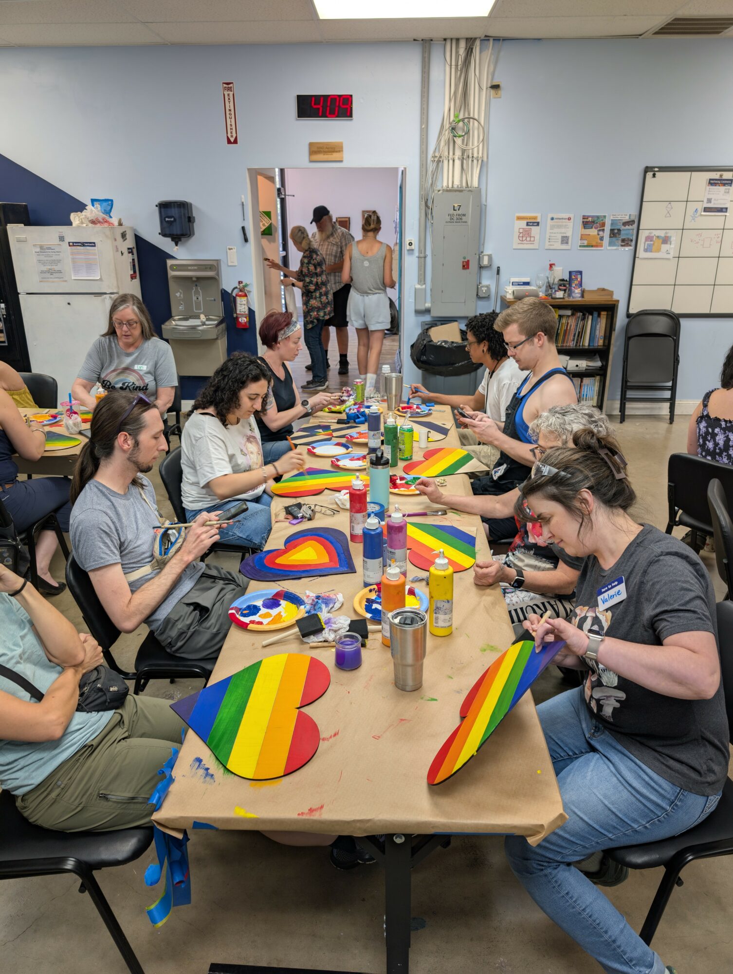 People sitting around a table painting rainbow-colored objects in a classroom with supplies and a whiteboard.