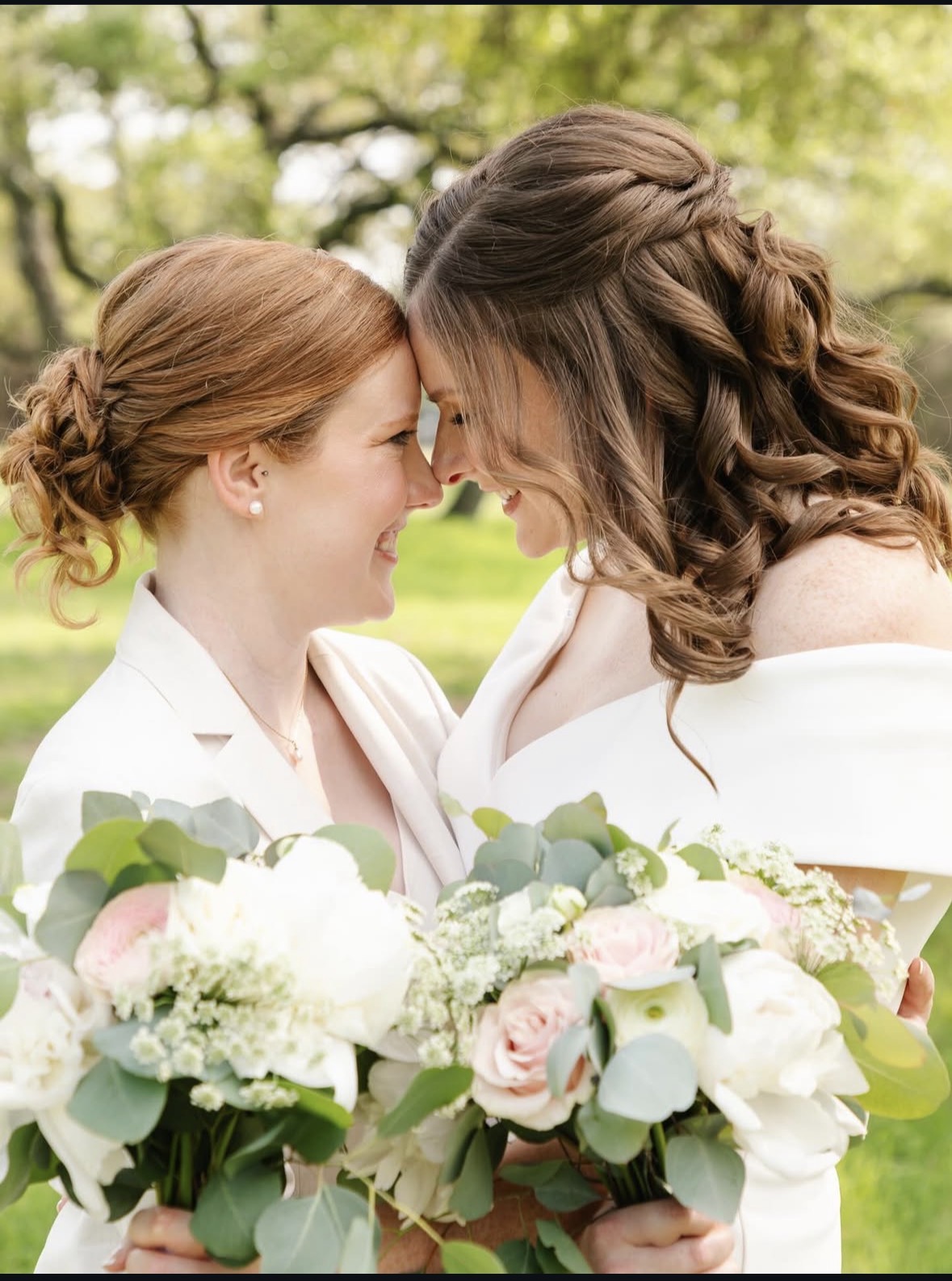 Two women face each other, smiling, holding a bouquet of white and pink flowers outdoors with trees in the background.