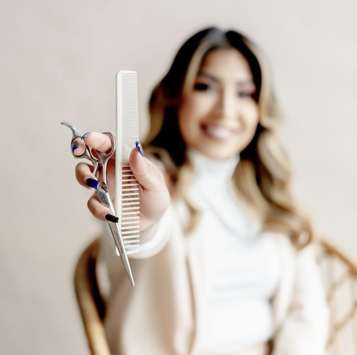 Person holding scissors, comb, and hairbrush, smiling, with blurred background.