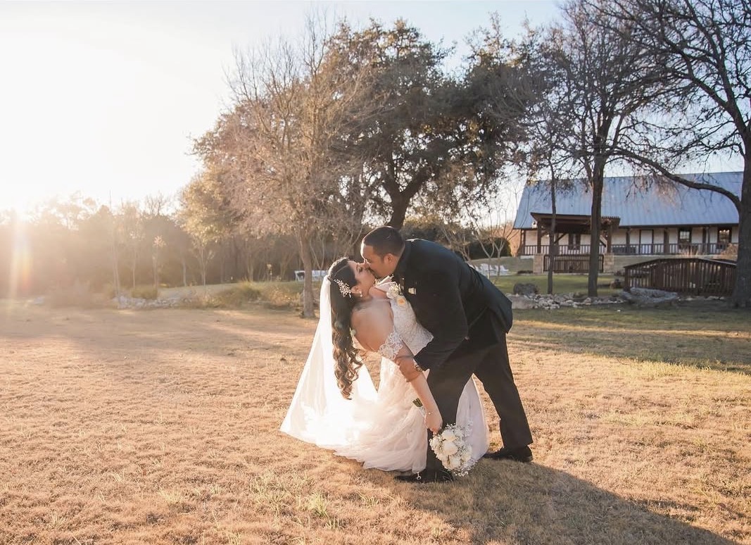 A couple in wedding attire sharing a kiss outdoors during sunset, with trees and a building in the background.
