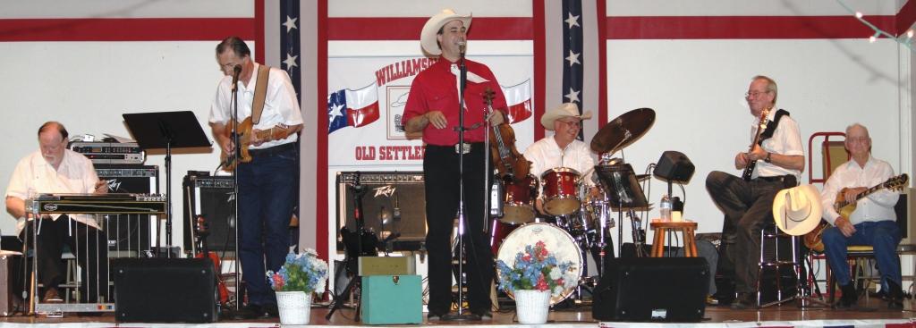 Group of six musicians performing on stage with American flags in background, playing various instruments, including drums, guitar, and keyboard.