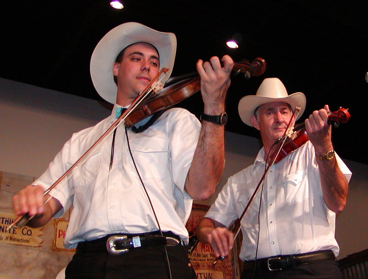 Two men wearing white shirts and cowboy hats playing violins on stage.