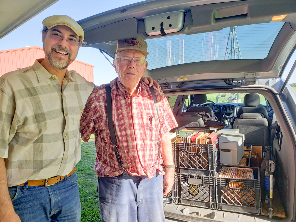 Two men standing next to an open vehicle trunk filled with boxes and crates, outdoors with a building in the background.
