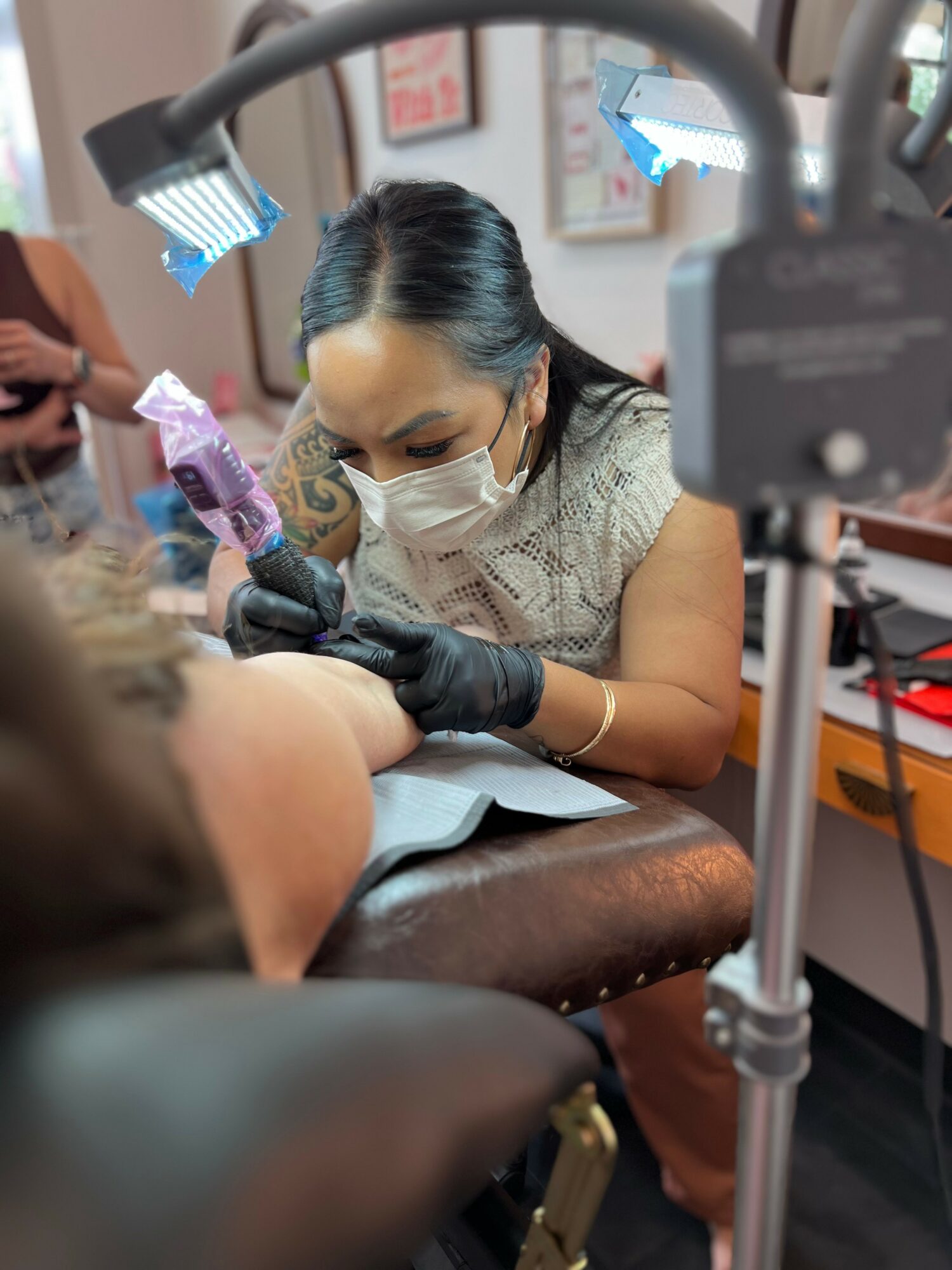 A woman wearing a face mask and gloves is administering a tattoo or medical procedure to a person's arm, with bright lights overhead.