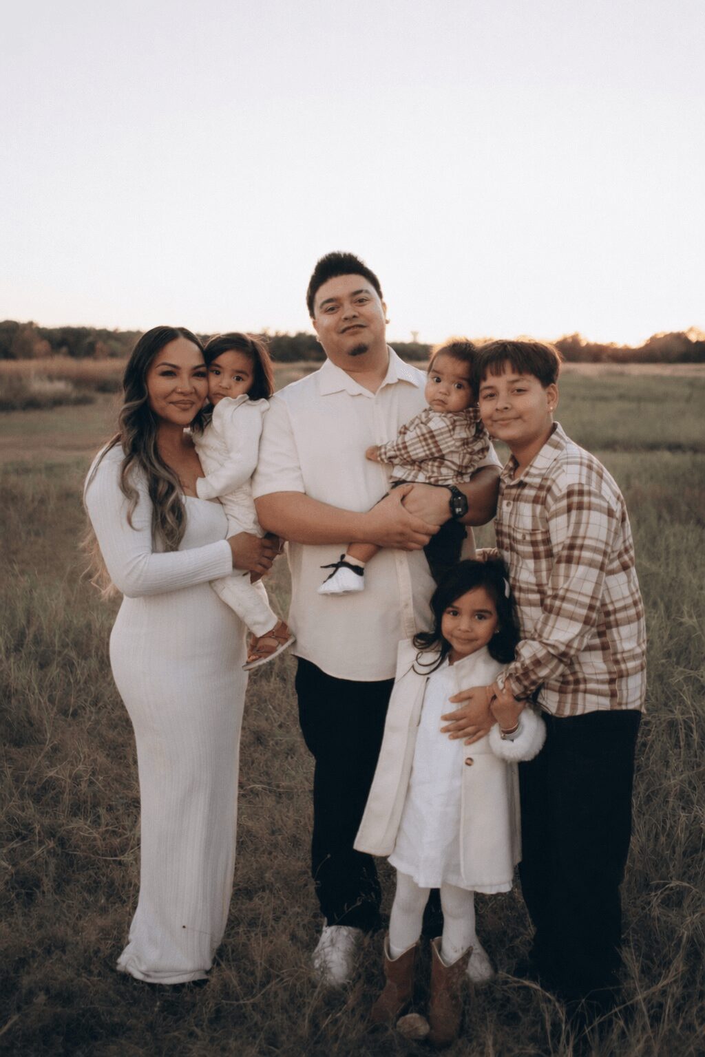 Family of six standing outdoors in a field, smiling, with a clear sky background.