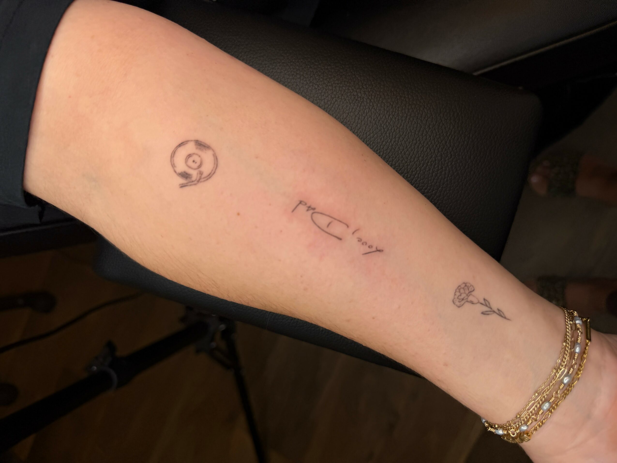 Tattooed forearm with spiral, text, and small symbol, wearing bracelets, against a dark background.