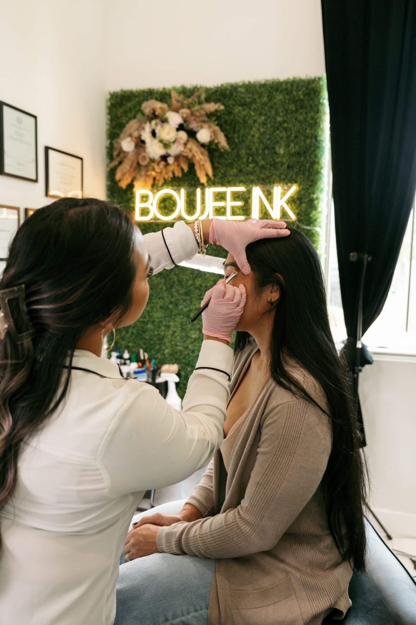 Makeup artist applying makeup to a woman seated indoors with a green wall and illuminated sign in the background.