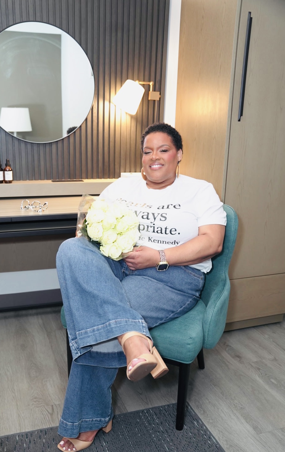 Woman sitting in a teal chair holding a bouquet of white flowers, smiling, in a modern room with a round mirror and lamp.