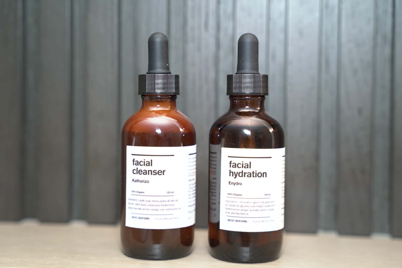 Two brown bottles with dropper caps labeled facial cleanser and facial hydration, placed on a surface against a metal background.