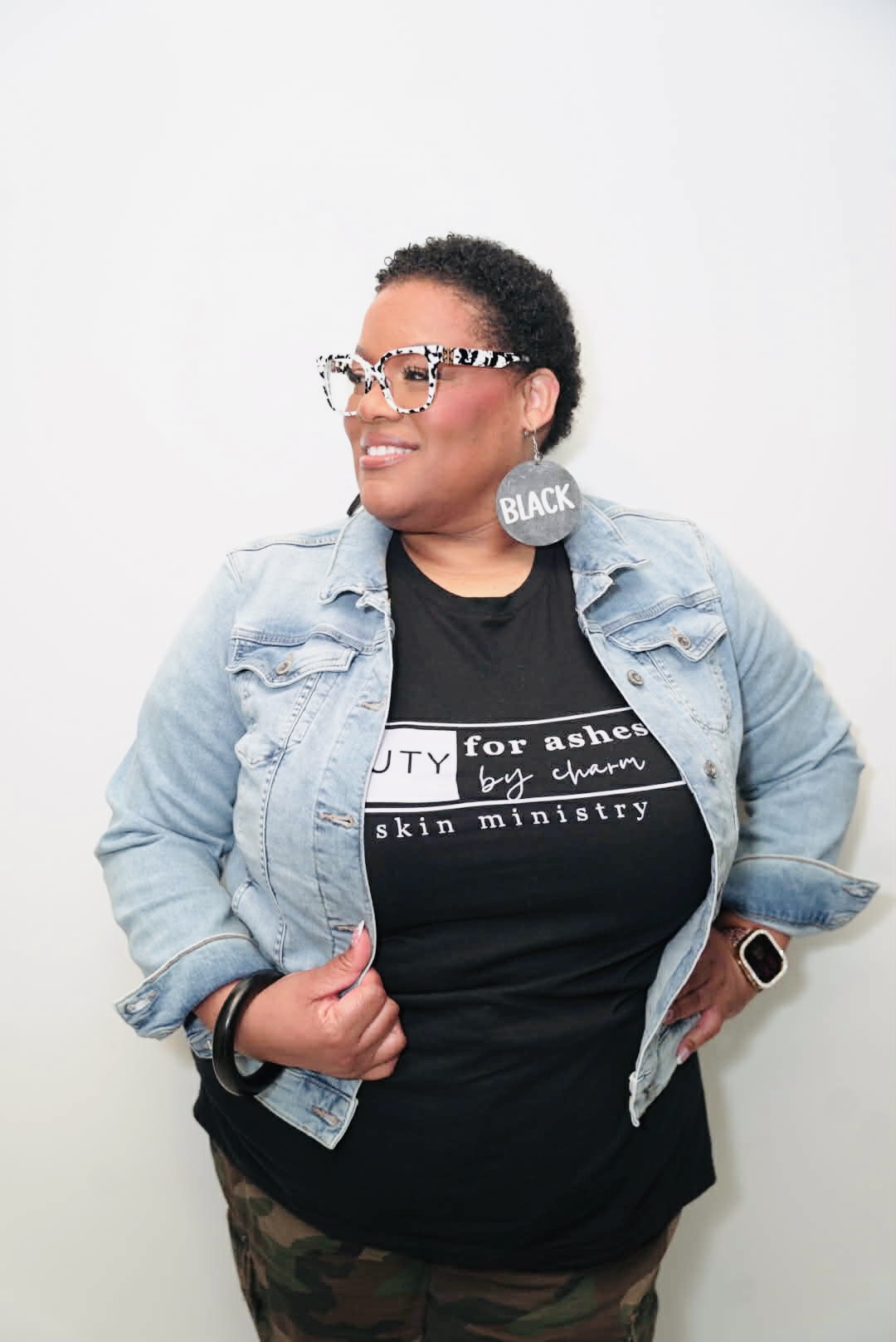Woman with short curly hair wearing glasses, denim jacket, black t-shirt, and camouflage pants, smiling and looking to the side.