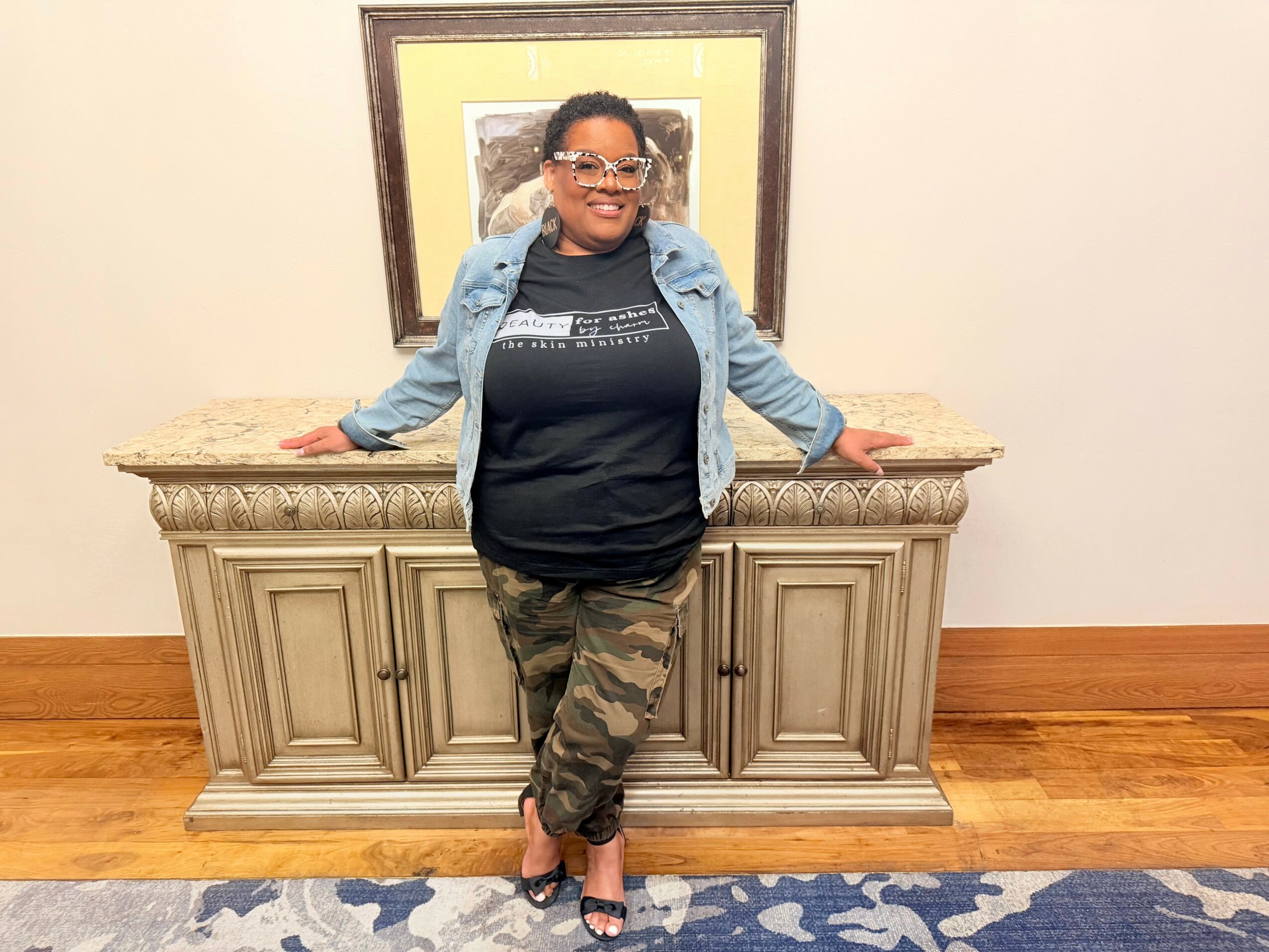 Woman standing in front of a decorative cabinet, smiling, wearing glasses, a denim jacket, black t-shirt, camouflage pants, and heels.