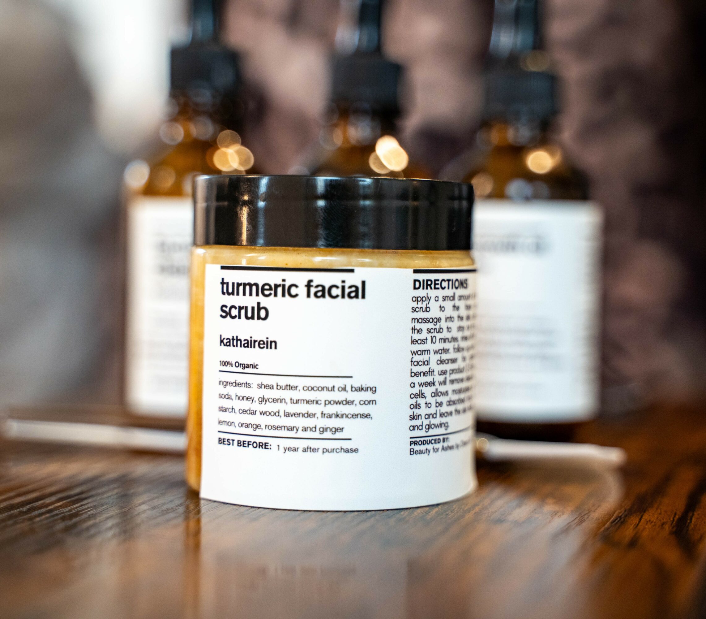 Container labeled turmeric facial scrub on a wooden surface with blurred bottles in background.