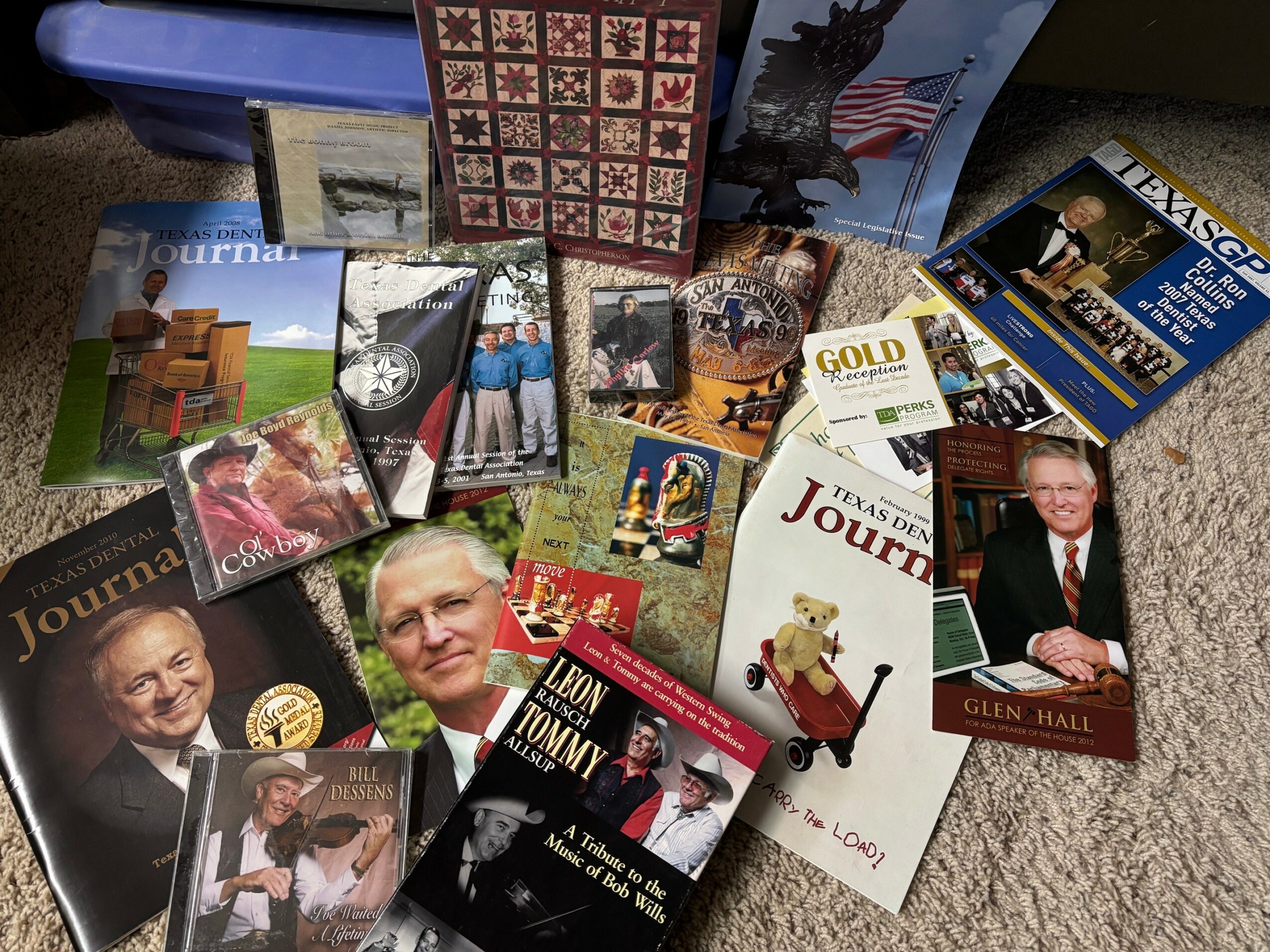 Collection of books, photographs, and memorabilia on a surface, including a portrait of a man, a teddy bear, and a quilt pattern.
