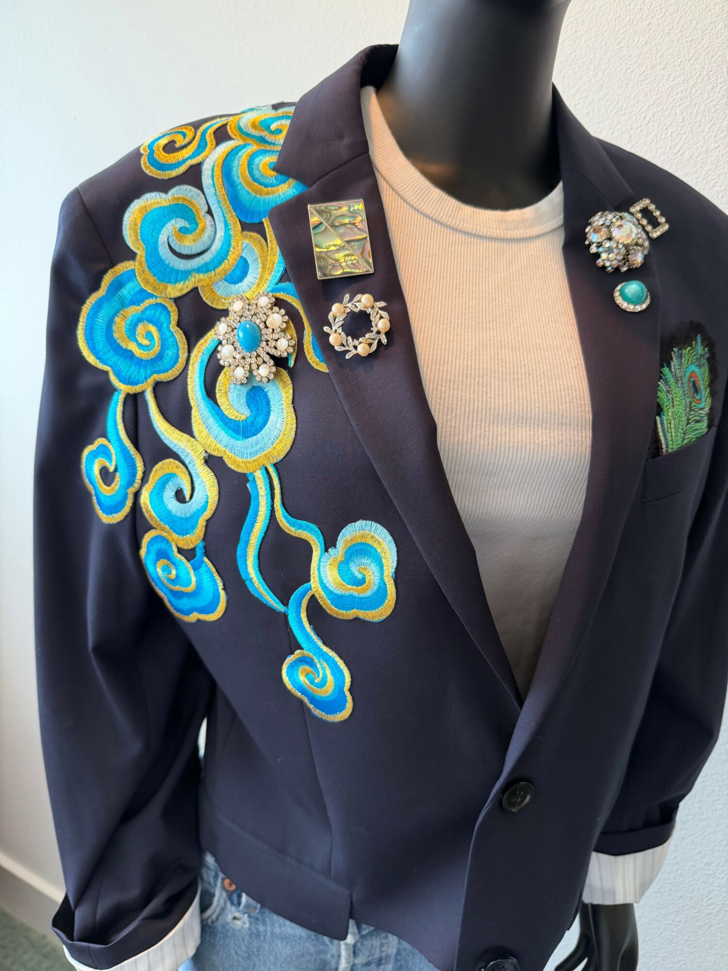 Black blazer with colorful swirling embroidery on the front, displayed on a mannequin torso, with various pins attached.