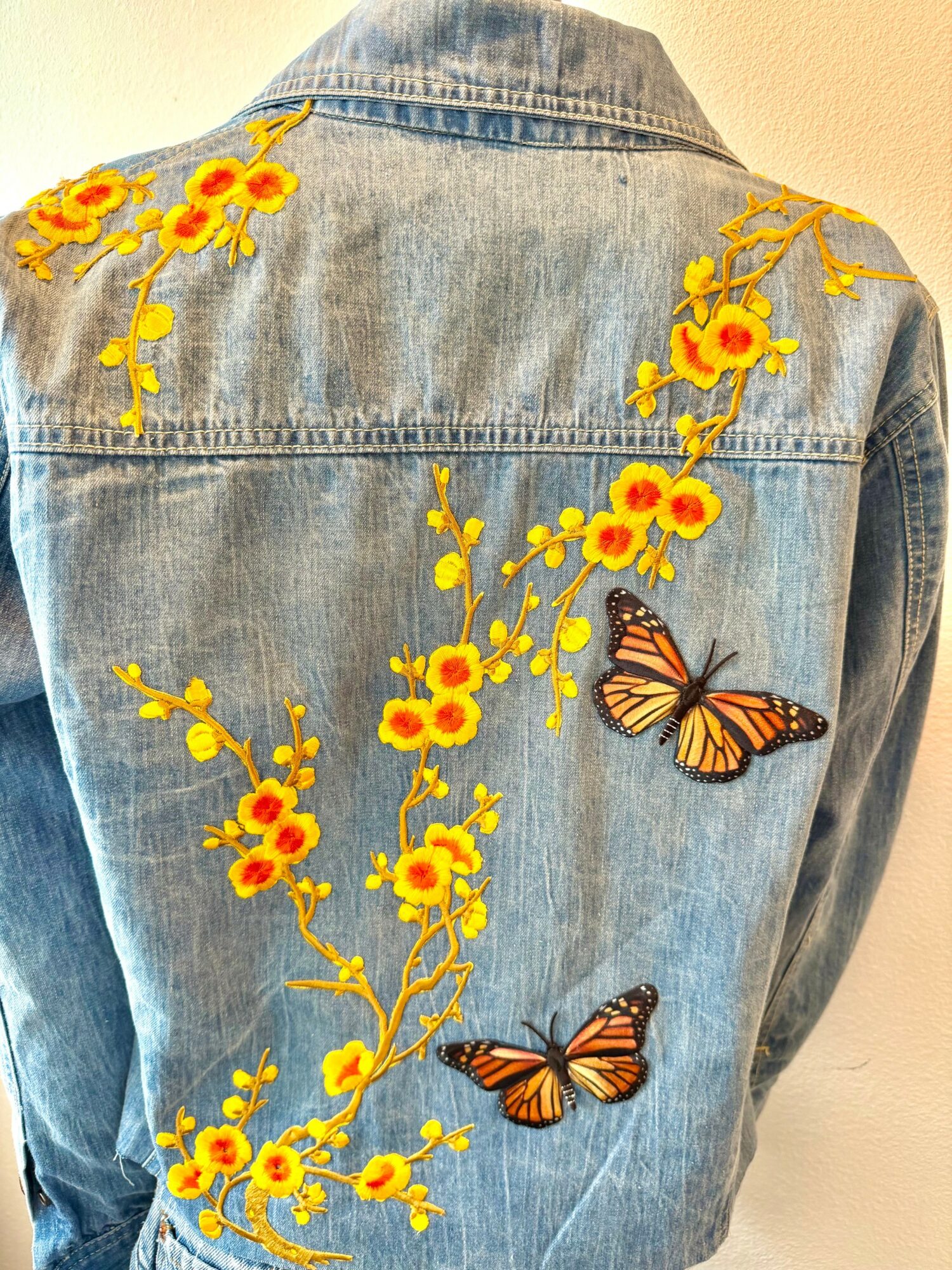 Back of denim jacket with yellow floral embroidery and two butterflies, one near the middle and one lower right.