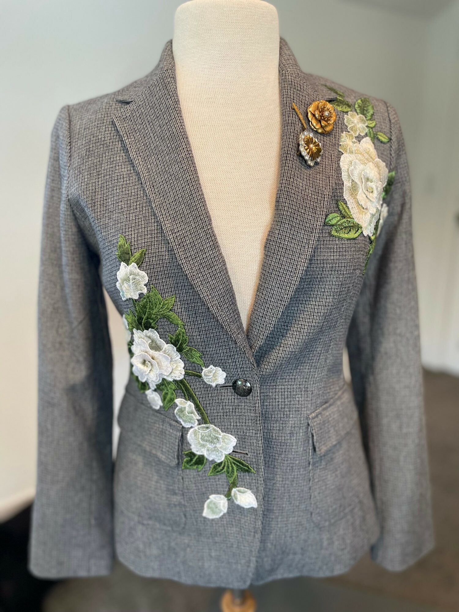 Gray blazer with floral embroidery on the left shoulder and right side, featuring white, yellow, and green flowers, and a single button.