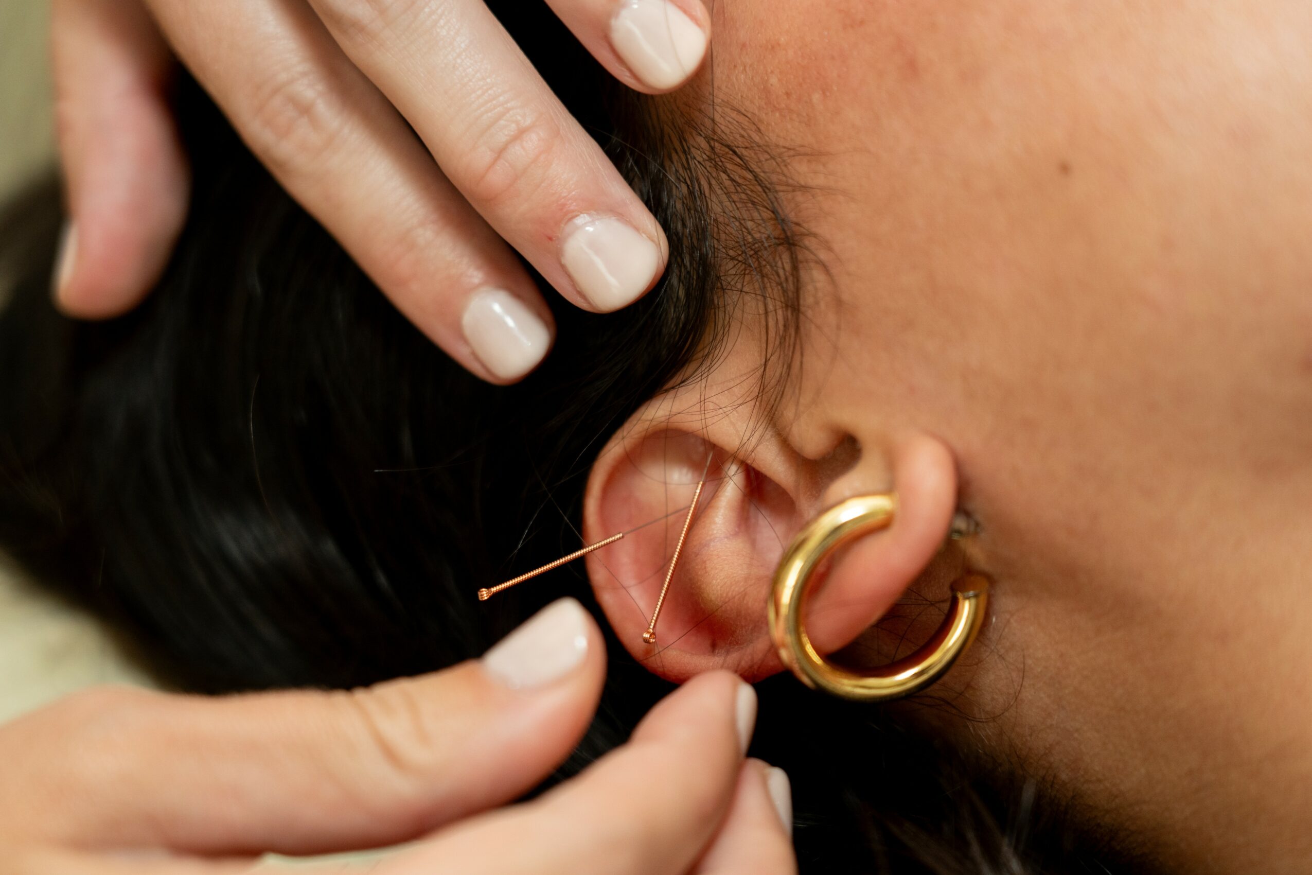 Close-up of a person's ear with multiple earrings, including a large gold hoop and a small stud, with hands adjusting earrings.