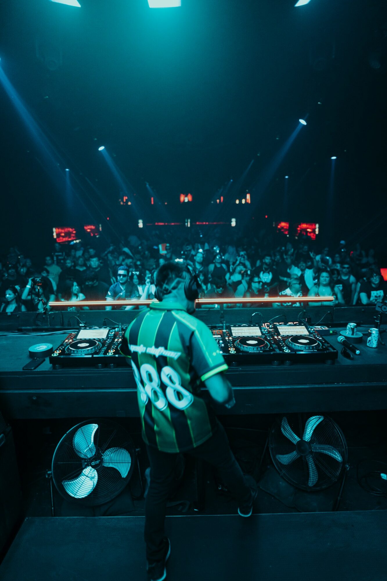 DJ stands at turntables in a dark club with a large crowd and blue lighting.
